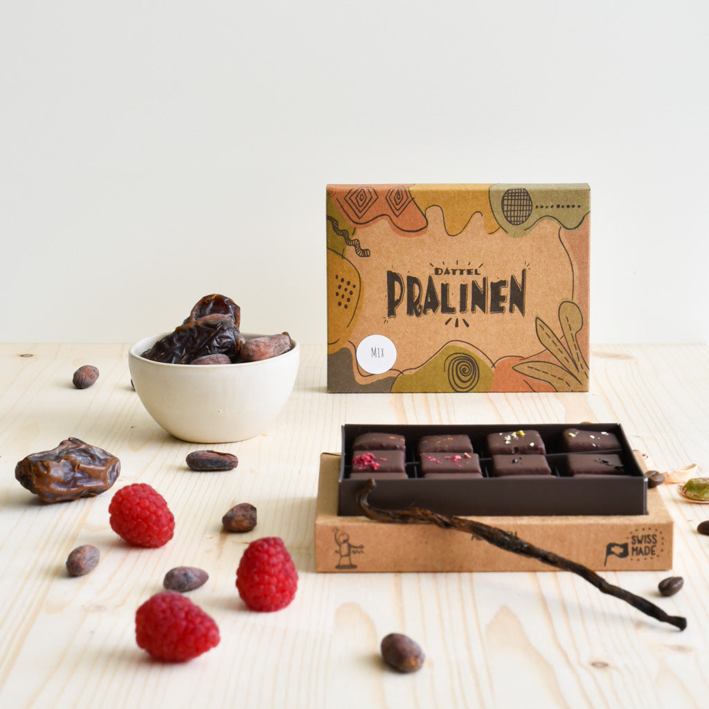 12er Pralinenbox frontal – Schachtel mit handgemachten Dattel-Pralinen, Zutaten und Schokolade mit Kokosblütenzucker im Vordergrund