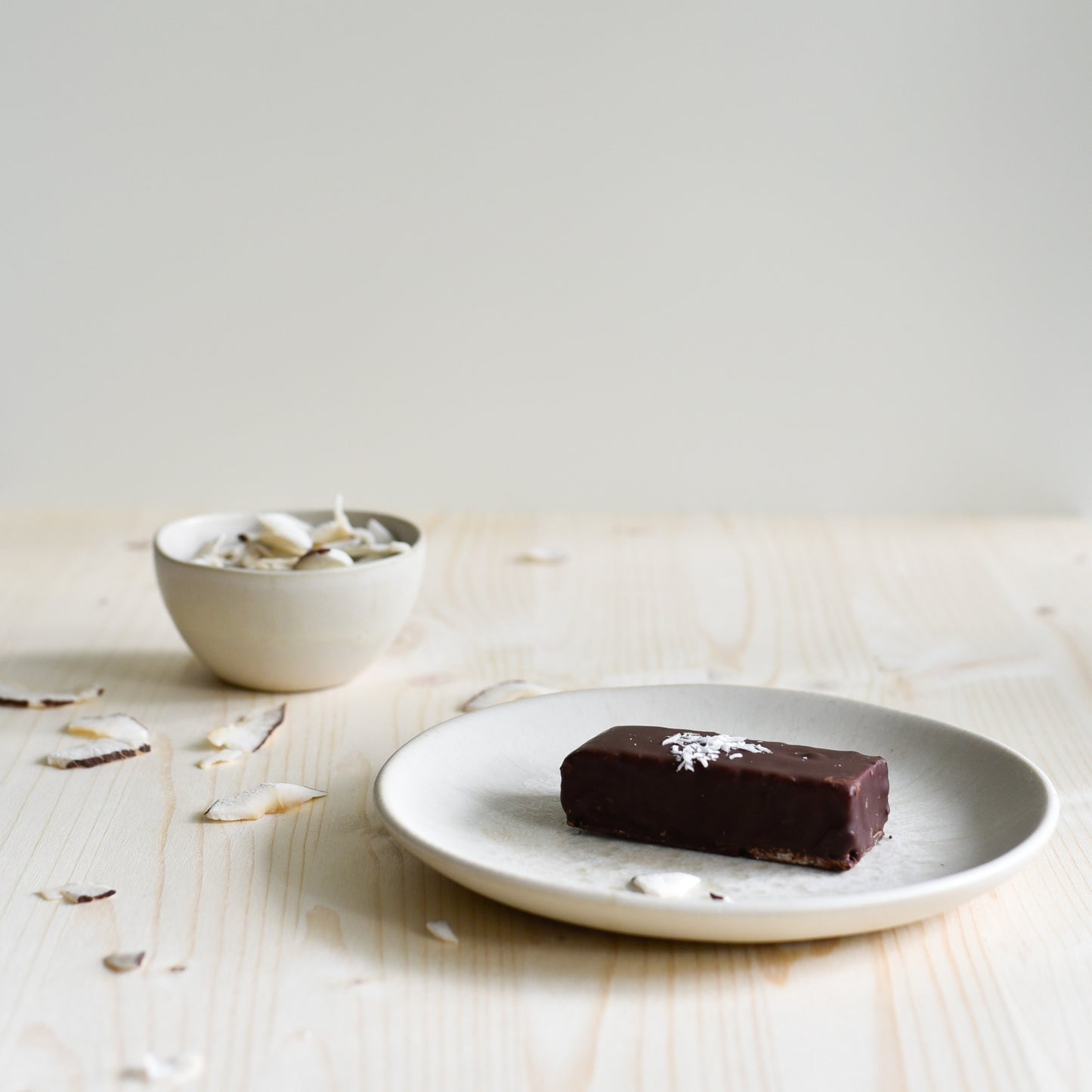 Nounty Kokosriegel ausgepackt, dunkle Schokolade sichtbar, Zutaten Datteln und Kokos im Hintergrund