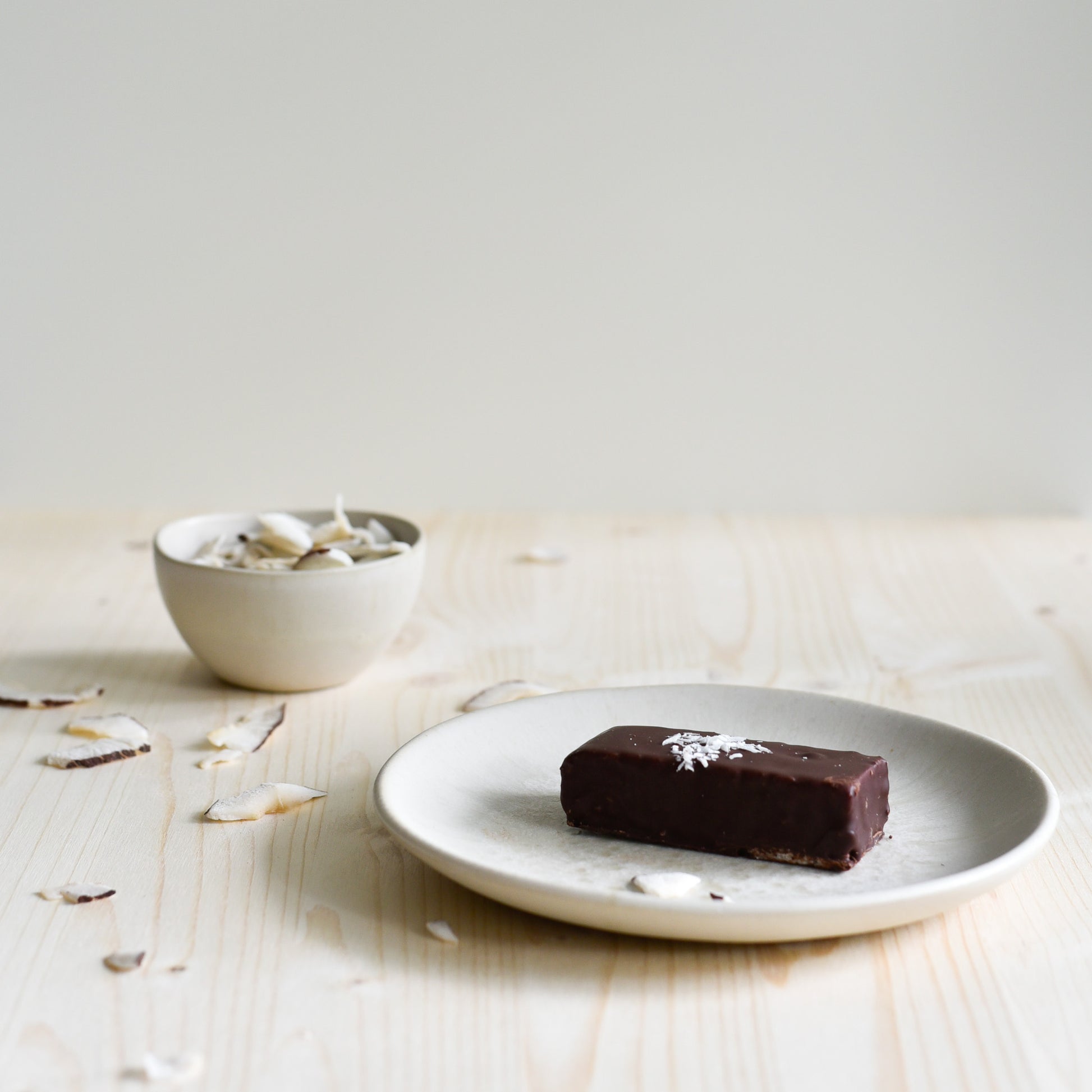 Nounty Kokosriegel ausgepackt, dunkle Schokolade sichtbar, Zutaten Datteln und Kokos im Hintergrund